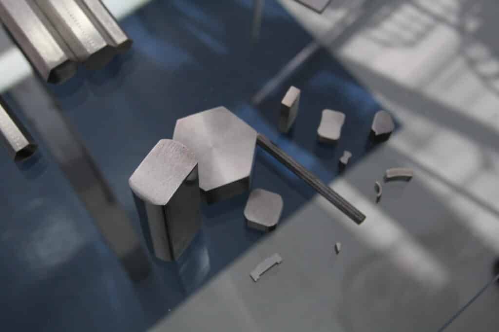 Um close-up mostra diversas peças e perfis de aço inoxidável em diferentes formatos (hexagonais, retangulares e angulares) dispostas sobre uma superfície reflexiva escura, com algumas barras alongadas desfocadas ao fundo.